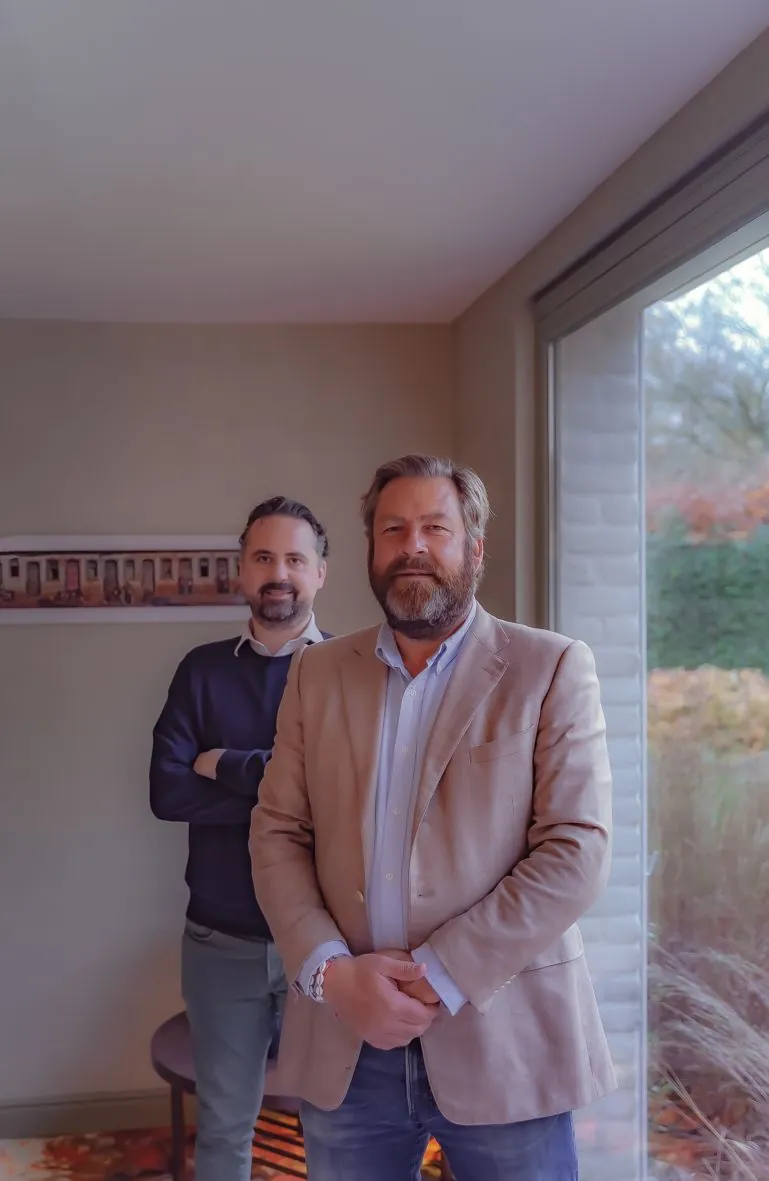 Deux hommes debout près d'une fenêtre, l'un en blazer beige, l'autre en tenue sombre